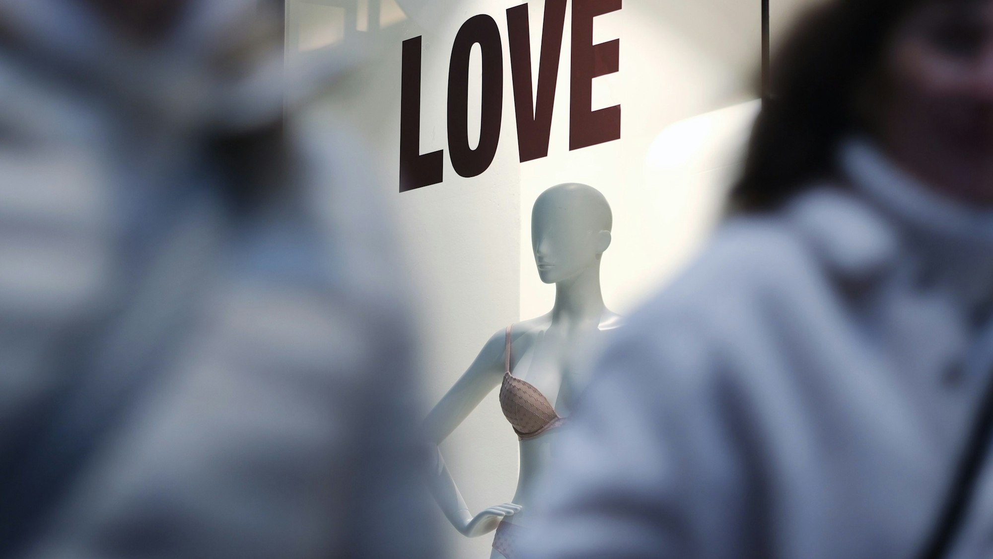 Man sieht zwischen zwei unscharfen Menschen durch fotografiert ein Schaufenster mit einer Puppe. Über ihr steht „LOVE“.