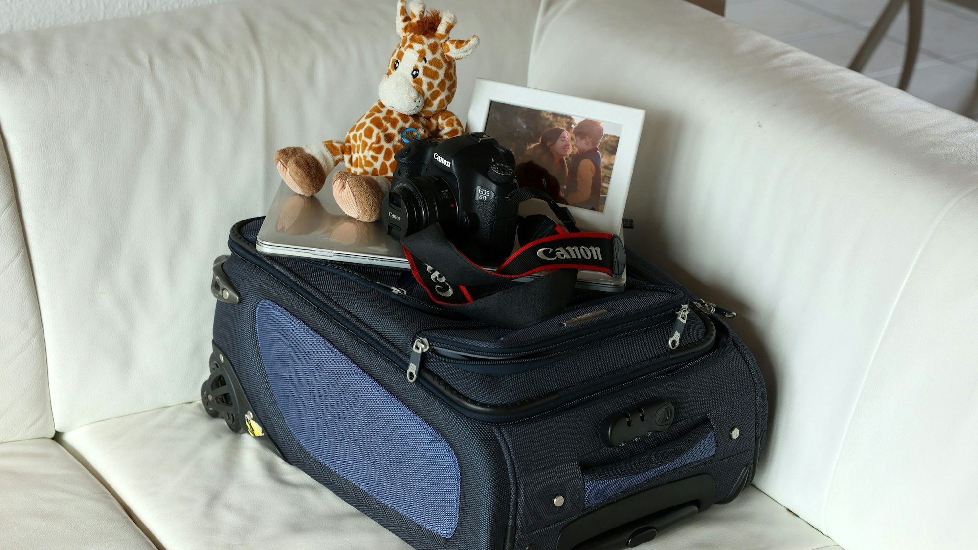 Auf einem weißen Sofa liegt ein kleiner, blauer Koffer. Darauf liegen eine Stoffgiraffe, ein Laptop, eine Kamera und ein gerahmtes Bild von Olga und ihrem Sohn.