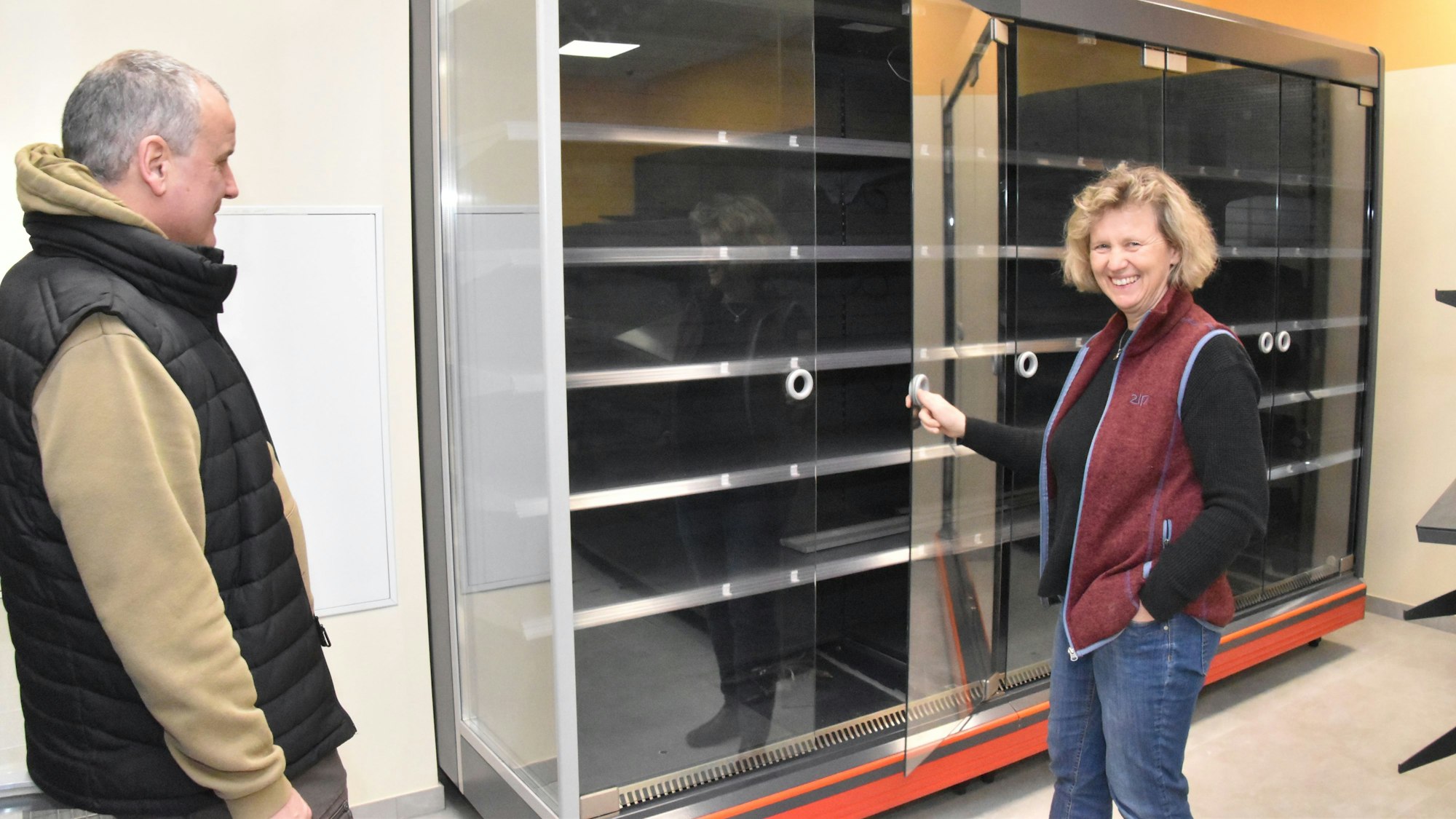 Zwei Personen stehen vor einem Warenregal, das noch leer ist. Ute Klein lächelt in die Kamera.