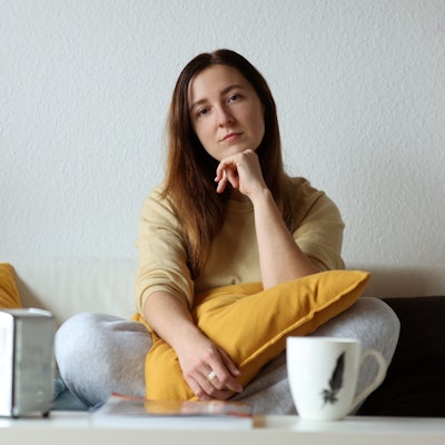 Olga sitzt im Schneidersitz auf dem Sofa in ihrer Wohnung. Sie guckt in die Kamera und hat ihren Ellenbogen auf ein gelbes Kissen gestützt, das auf ihrem Schoß liegt.