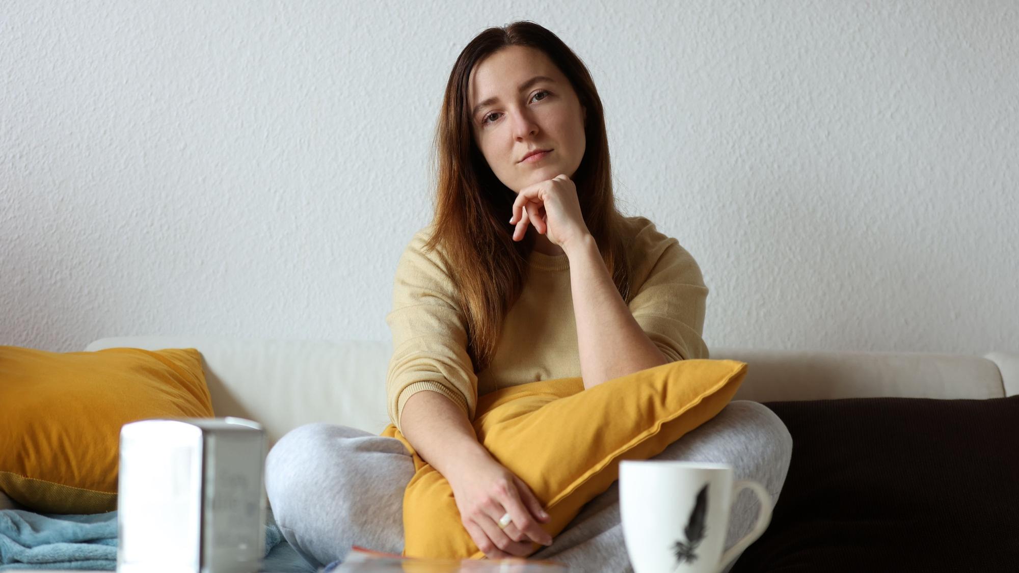 Olga sitzt im Schneidersitz auf dem Sofa in ihrer Wohnung. Sie guckt in die Kamera und hat ihren Ellenbogen auf ein gelbes Kissen gestützt, das auf ihrem Schoß liegt.