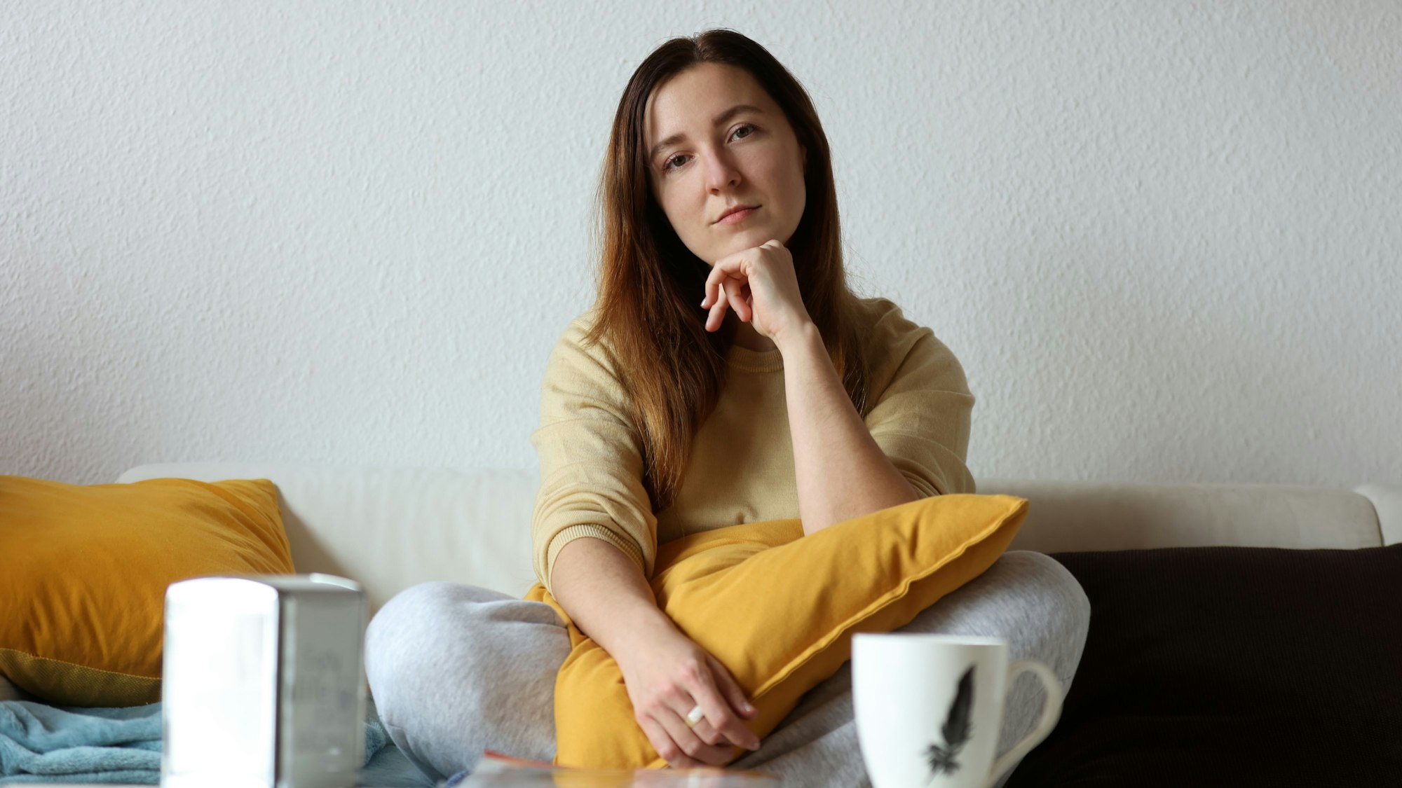 Olga sitzt im Schneidersitz auf dem Sofa in ihrer Wohnung. Sie guckt in die Kamera und hat ihren Ellenbogen auf ein gelbes Kissen gestützt, das auf ihrem Schoß liegt.