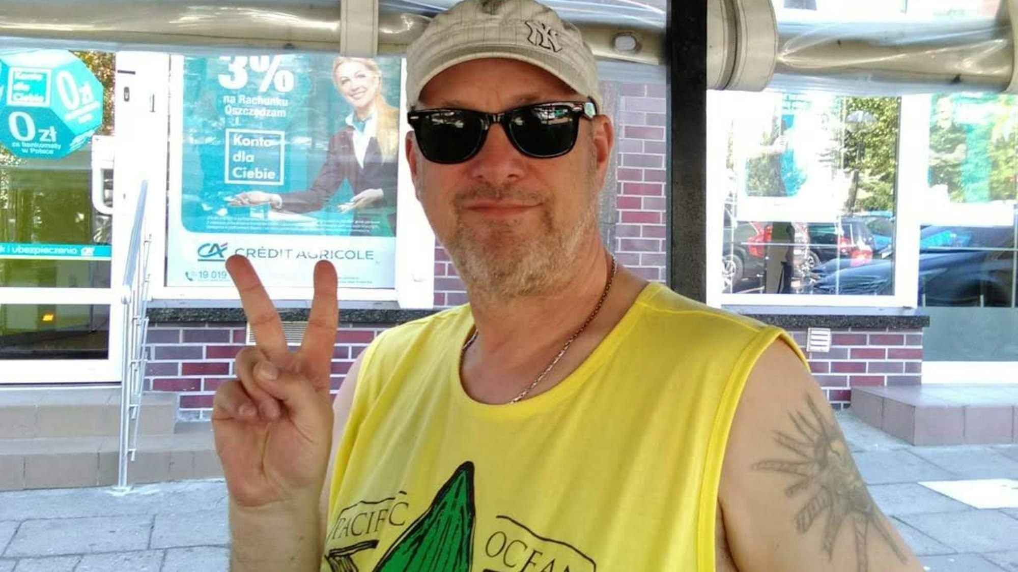 Michael K., mit Sonnenbrille und Käppie, trägt ein gelbes Shirt, lächelt in die Kamera und reckt Zeige- und Mittelfinger zu einem Peacezeichen in die Luft.