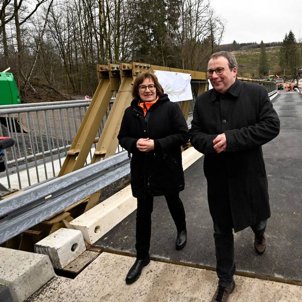 Oliver Krischer (Bündnis 90/Die Grünen), Verkehrsminister von Nordrhein-Westfalen, schaut sich neben Petra Beckefeld, Technische Direktorin des Landesbetriebs Straßenbau Nordrhein-Westfalen, eine defekte Brücke über der Wipper bei Wipperfürth samt Behelfsbau an. Beide tragen schwarze Jacken.