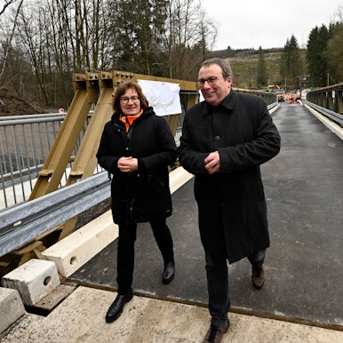 Oliver Krischer (Bündnis 90/Die Grünen), Verkehrsminister von Nordrhein-Westfalen, schaut sich neben Petra Beckefeld, Technische Direktorin des Landesbetriebs Straßenbau Nordrhein-Westfalen, eine defekte Brücke über der Wipper bei Wipperfürth samt Behelfsbau an. Beide tragen schwarze Jacken.