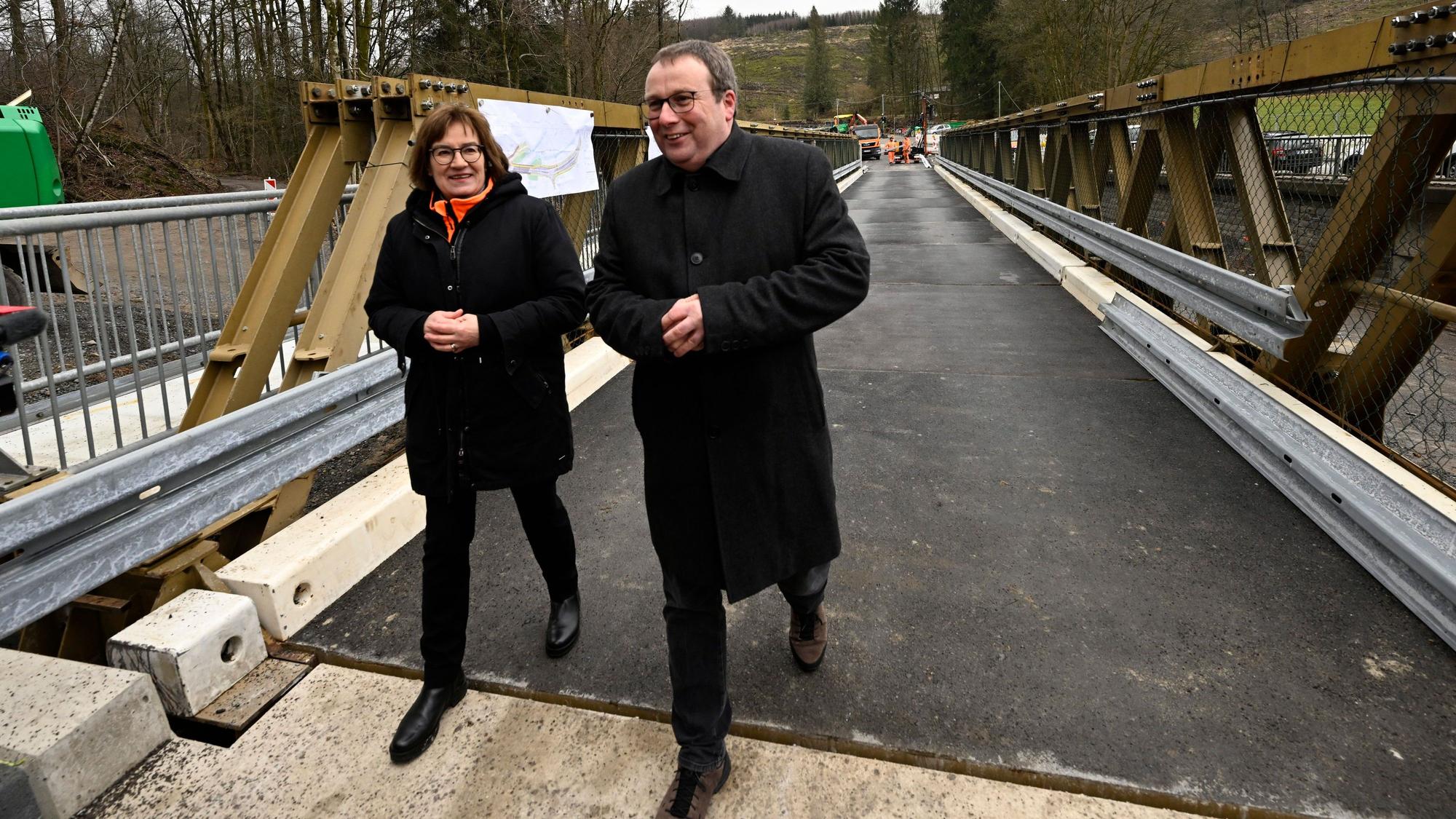 Oliver Krischer (Bündnis 90/Die Grünen), Verkehrsminister von Nordrhein-Westfalen, schaut sich neben Petra Beckefeld, Technische Direktorin des Landesbetriebs Straßenbau Nordrhein-Westfalen, eine defekte Brücke über der Wipper bei Wipperfürth samt Behelfsbau an. Beide tragen schwarze Jacken.