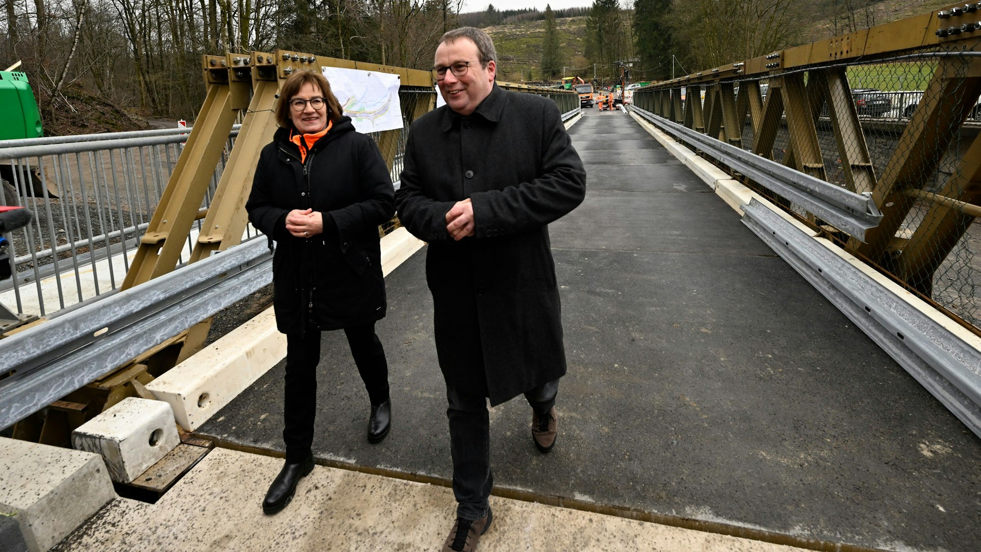 Oliver Krischer (Bündnis 90/Die Grünen), Verkehrsminister von Nordrhein-Westfalen, schaut sich neben Petra Beckefeld, Technische Direktorin des Landesbetriebs Straßenbau Nordrhein-Westfalen, eine defekte Brücke über der Wipper bei Wipperfürth samt Behelfsbau an. Beide tragen schwarze Jacken.