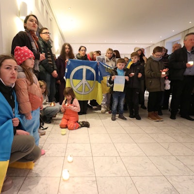 Eine Frau hat die blau-gelbe ukrainische Flagge umgehangen. Auf dem Boden stehen Windlichter. Im Hintergrund halten Menschen eine ukrainische Flagge mit dem Peace-Zeichen.