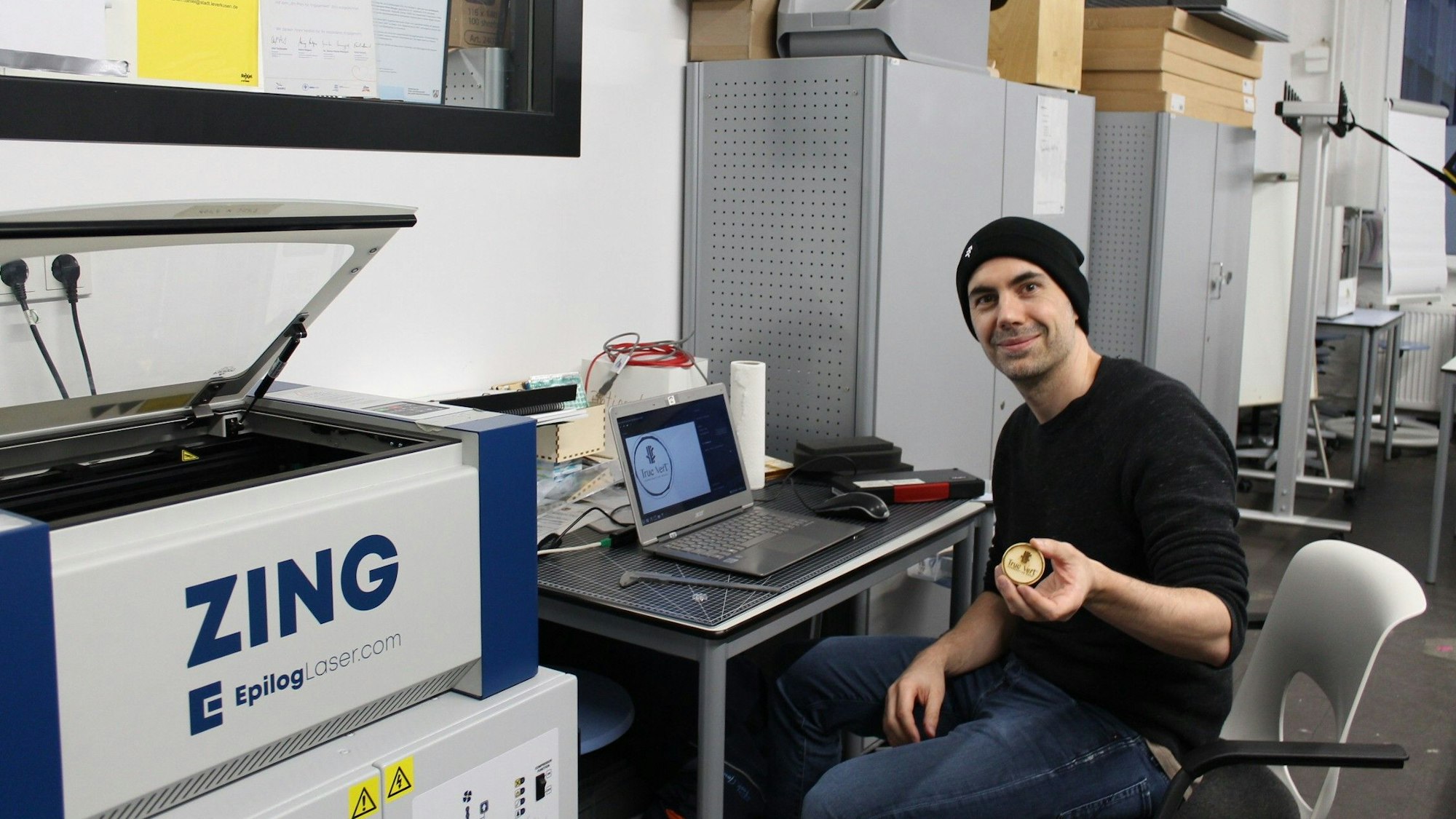 Benjamin Jürgens (40) ist Experte im Umgang mit dem Lasercutter. Auf dem Bild sitzt er vor einem Laptop am Schreibtisch und hält ein kleines, bearbeitetes Stück Holz in die Kamera.