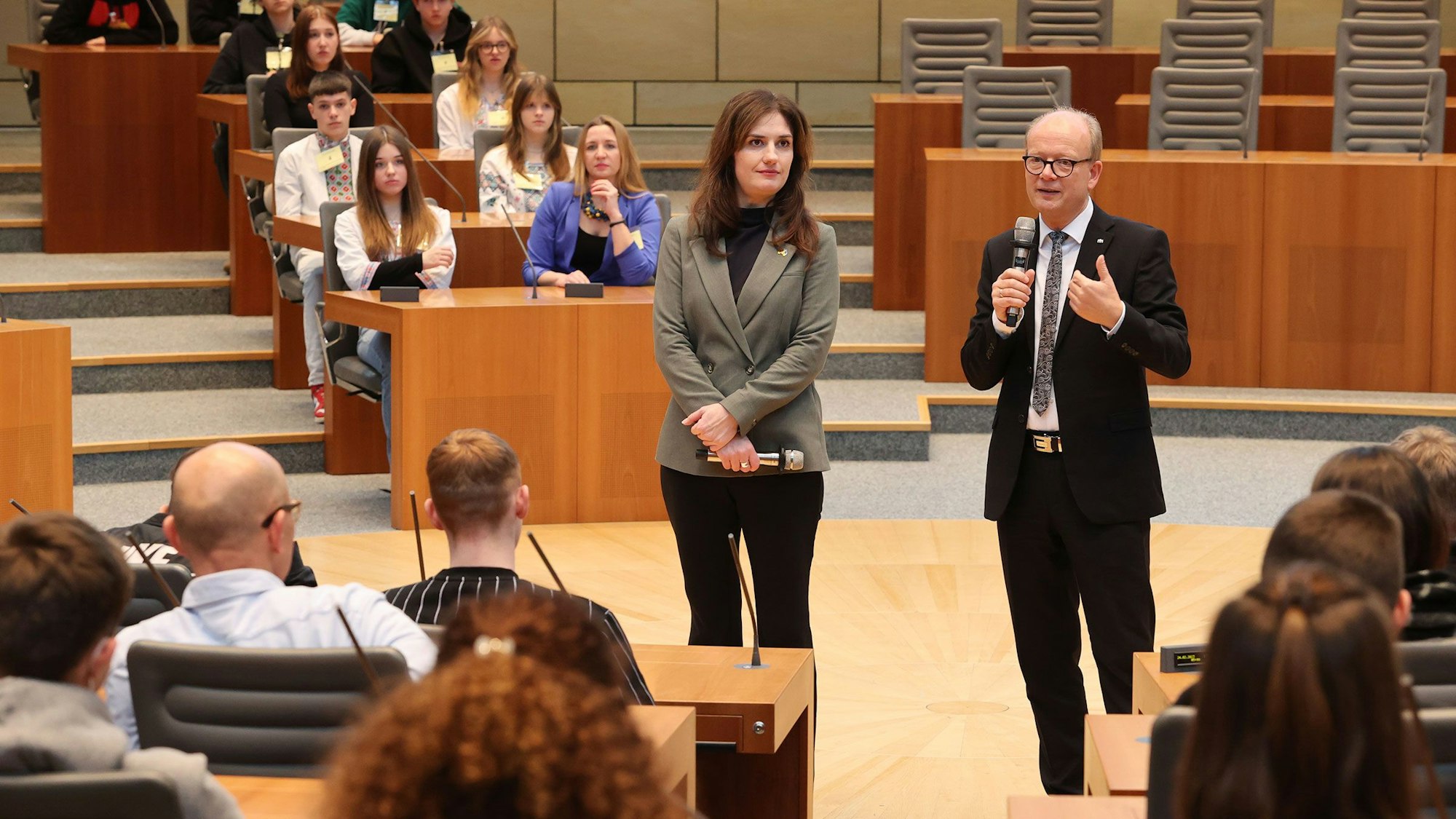 Iryna Shum und André Kuper stehen in der mitte des Plenarsaals, Kuper hat ein Mikrofon in der Hand und spricht. Auf den Plätzen der Abgeordneten sitzen die Schülerinnen und Schüler.