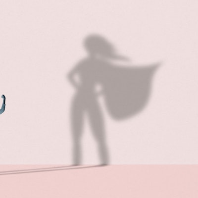 Eine junge Frau streckt ihre Arme in die Höhe, vor einer rosafarbenen Wand, auf der der Schatten einer Superwoman erscheint.