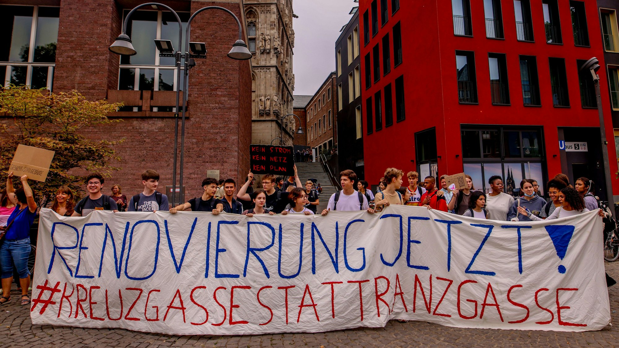Schüler und Eltern des Gymnasiums Kreuzgasse demonstrieren im Sommer 2022 für eine Sanierung des Schulgebäudes vor dem Historischen Rathaus auf dem Alter Markt in Köln. Sie halten ein Transparent mit den Worten „Renovierung Jetzt! Kreuzgasse statt Ranzgasse“ hoch.