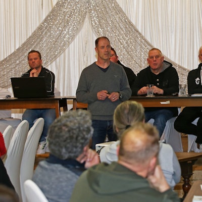 Mehrere Männer sitzen an einem Tisch. Einer steht im Vordergrund und hält einen Vortrag. Auf einer Leinwand ist eine Pärsentation zu sehen.