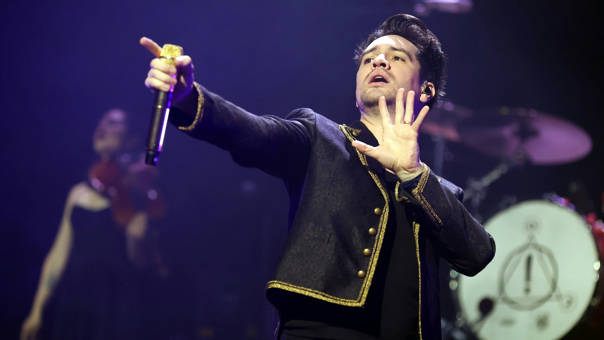 Urie trägt schwarze Jeans und Tshirt und ein schwarzes Sakko mit Goldverzierung.