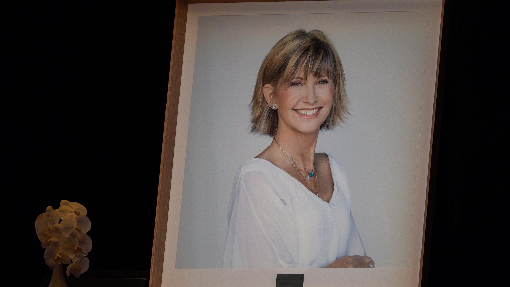 Porträt von Olivia Newton-John mit Blumen daneben.