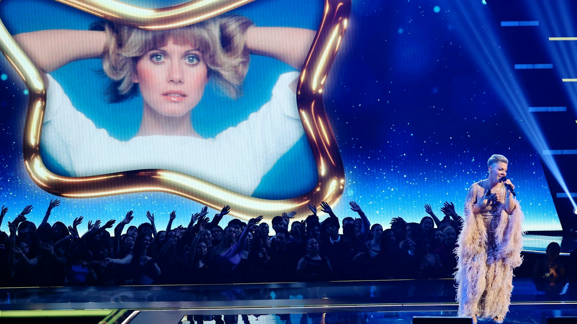 Pink singt auf der Bühne. Im Hintergrund ein Bild von Olivia Newton-John.