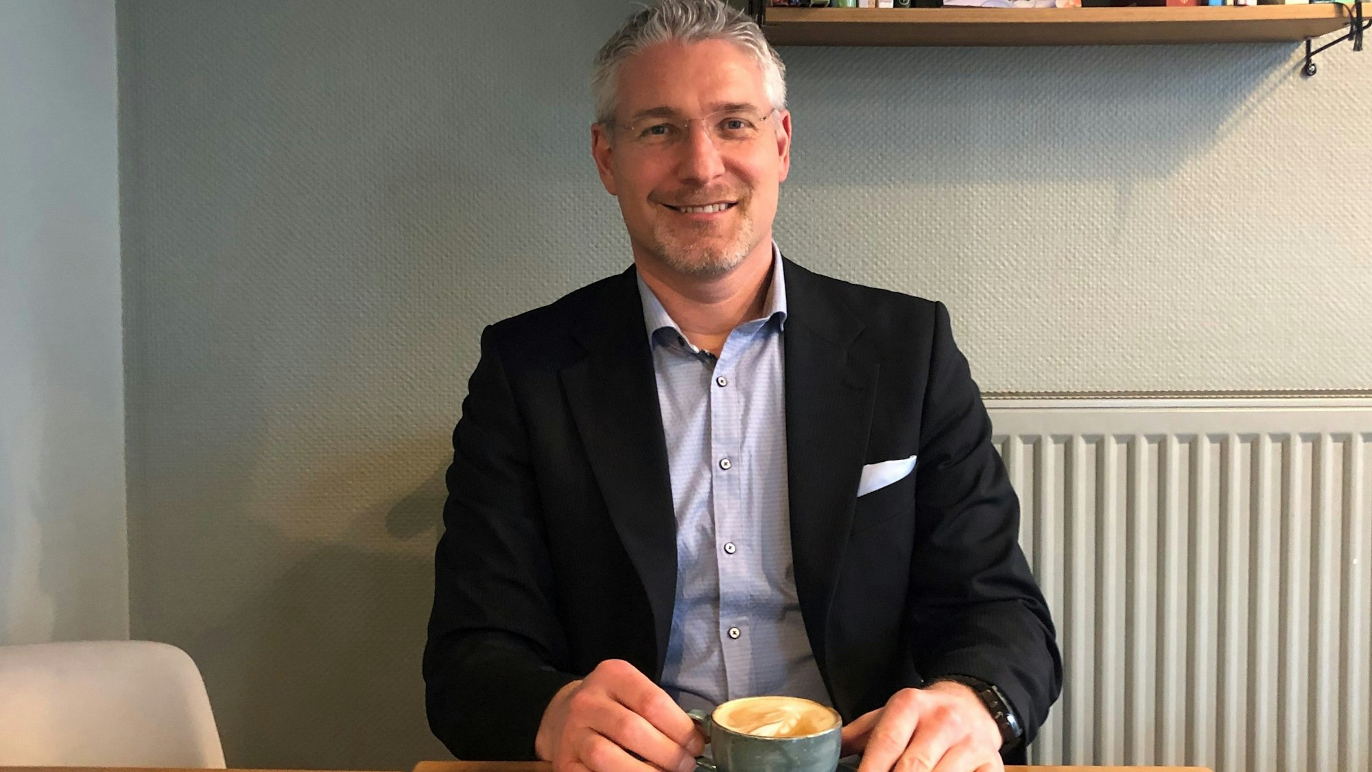 Martin Sagel aus Kerpen sitzt im Anzug in einem Café. In seinen Händen hält er einen Cappuccino.