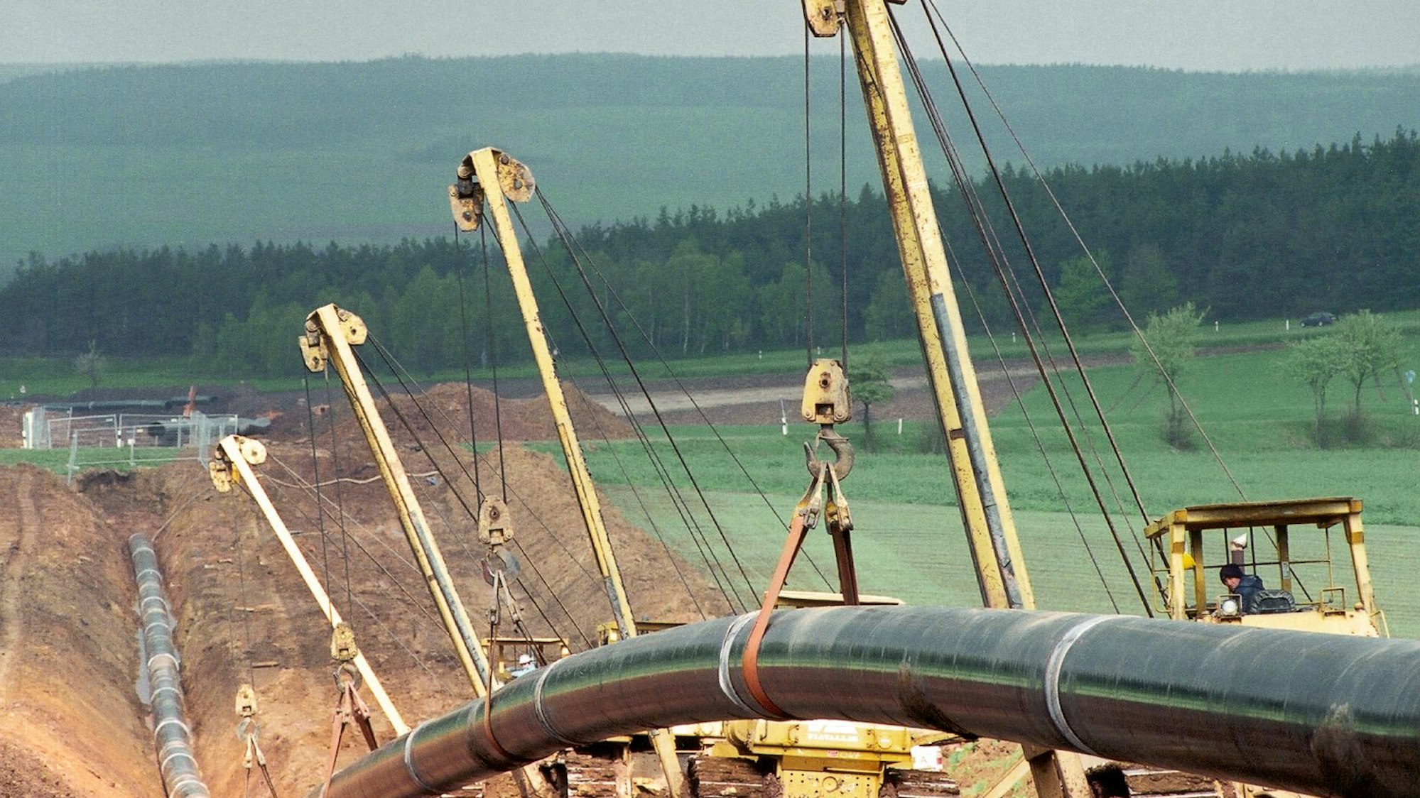 Silberne Rohre werden in riesige Furchen unter der Erde verlegt. Im Hintergrund steht ein Wald.