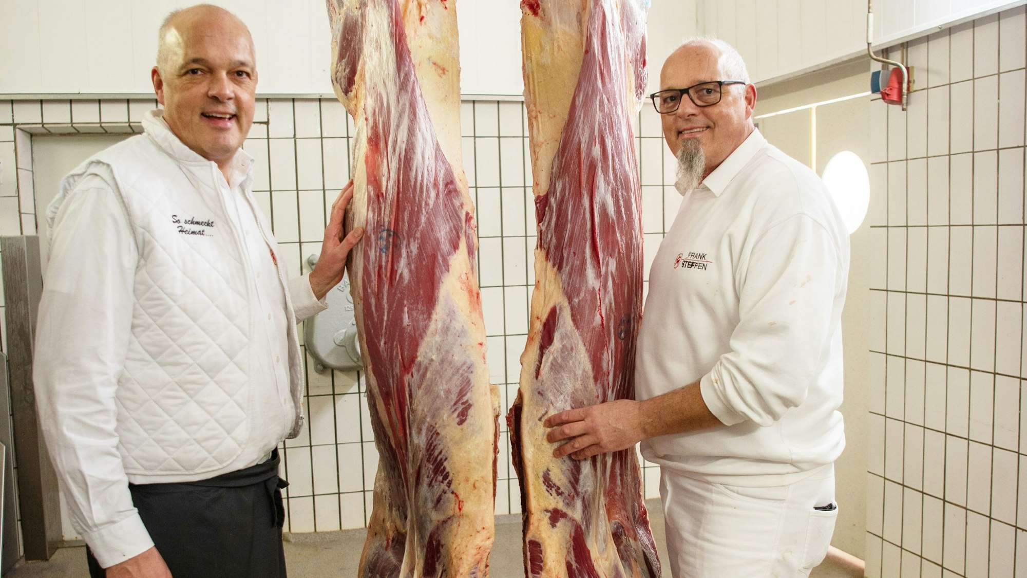 Zwei Männer stehen neben zwei aufgehängten Rinder-Vierteln.