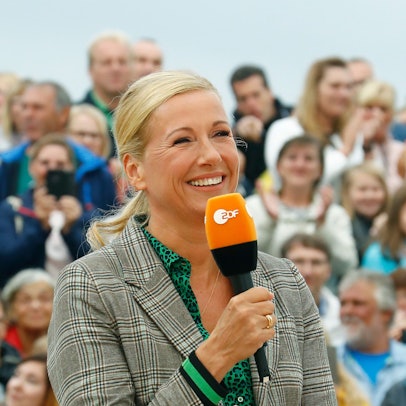 Andrea Kiewel (57) bei der Moderation des „ZDF-Fernsehgartens“. Die Moderatorin hält ein Mikrofon mit dem Senderlogo in der Hand, im Hintergrund ist das Publikum zu sehen.