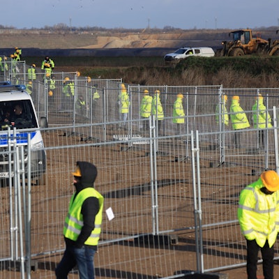 Mitarbeiter einer Werkschutzfirma stehen im Januar an einem Zaun beim von Klimaaktivisten besetzten Braunkohleort Lützerath. Sie alle tragen neongelbe Warnjacken. Hinter dem Zaun sind Vans der Polizei zu sehen.