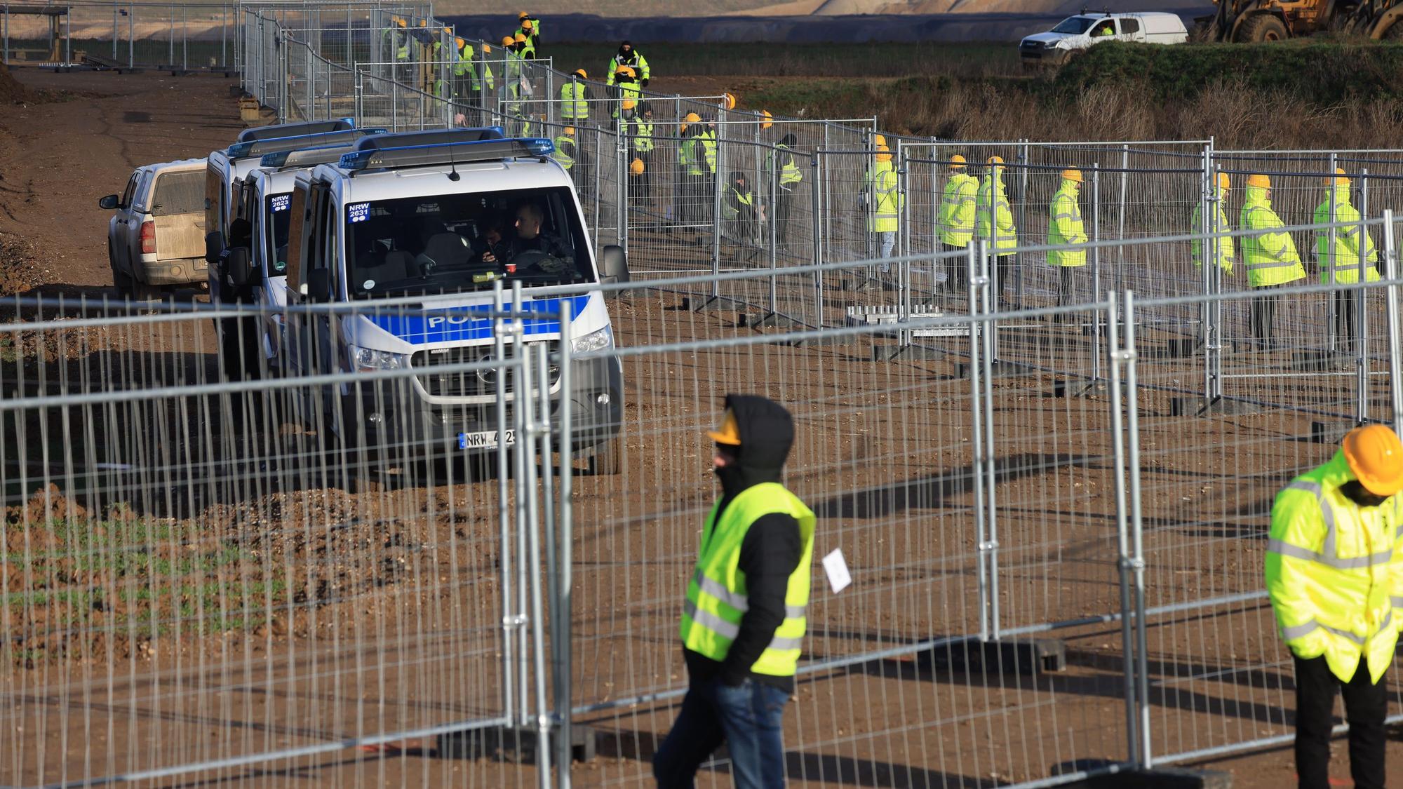 Mitarbeiter einer Werkschutzfirma stehen im Januar an einem Zaun beim von Klimaaktivisten besetzten Braunkohleort Lützerath. Sie alle tragen neongelbe Warnjacken. Hinter dem Zaun sind Vans der Polizei zu sehen.