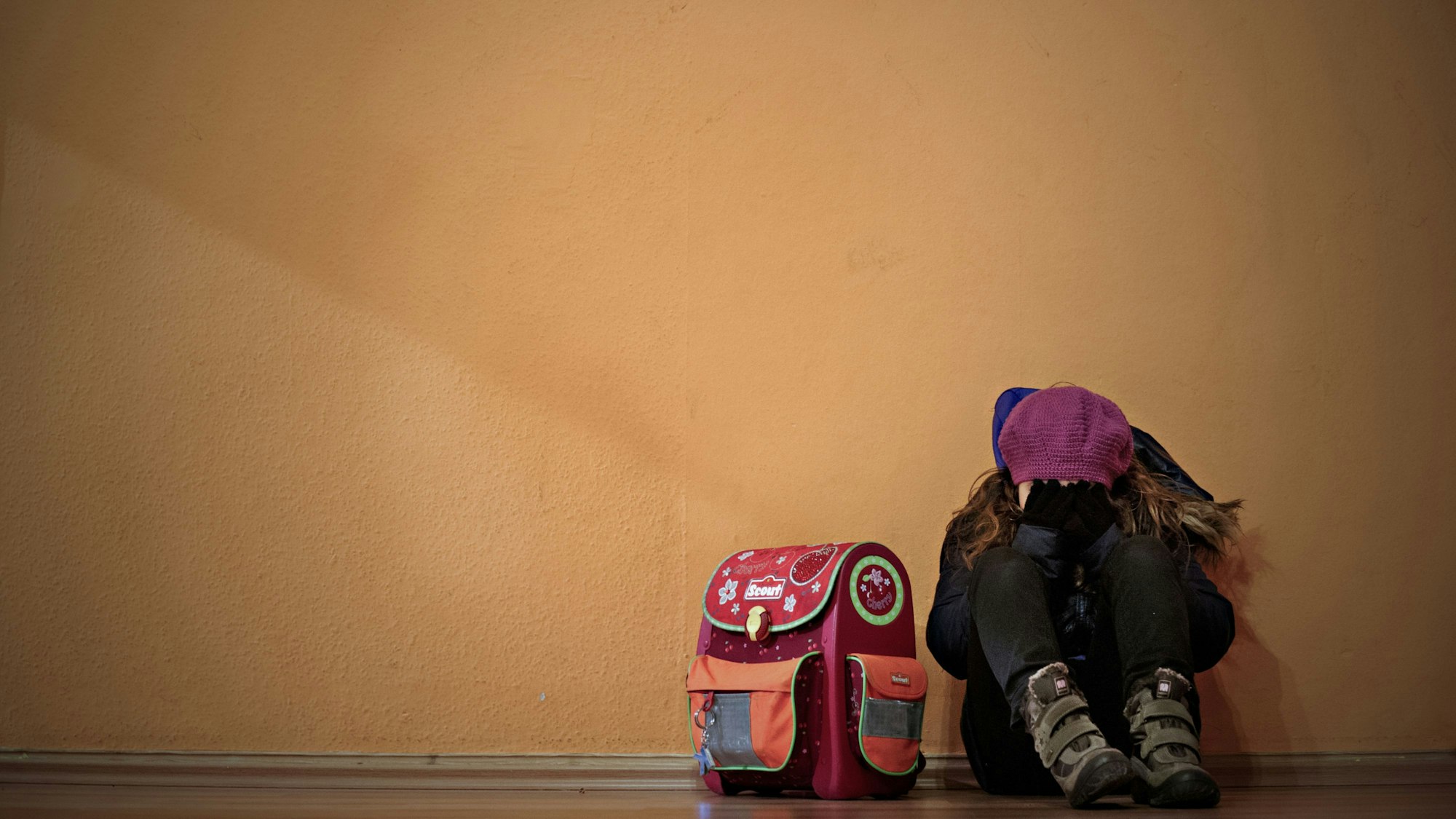 Ein Mädchen sitzt neben ihrem Schulranzen auf dem Boden und hält die Hände vor das Gesicht.