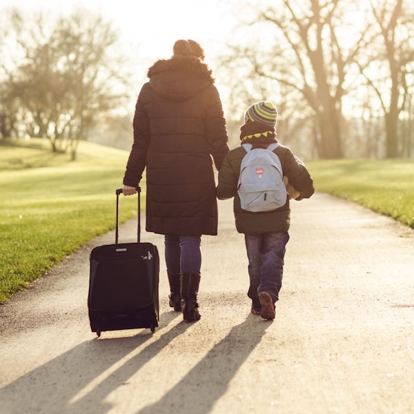Eine Frau geht mit einem Rollkoffer in der einen Hand und einem Sohn an der anderen Hand einen Weg entlang.