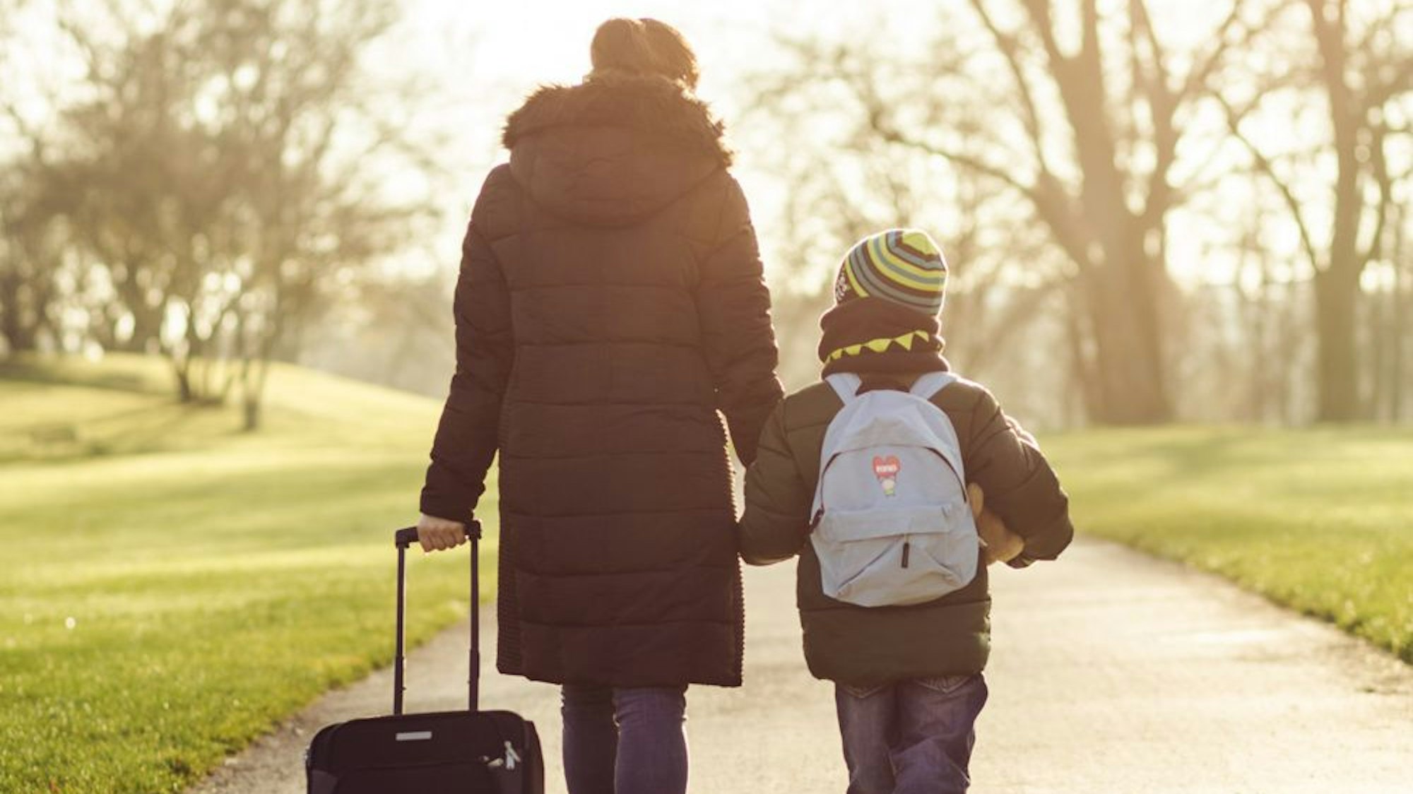 Eine Frau geht mit einem Rollkoffer in der einen Hand und einem Sohn an der anderen Hand einen Weg entlang.