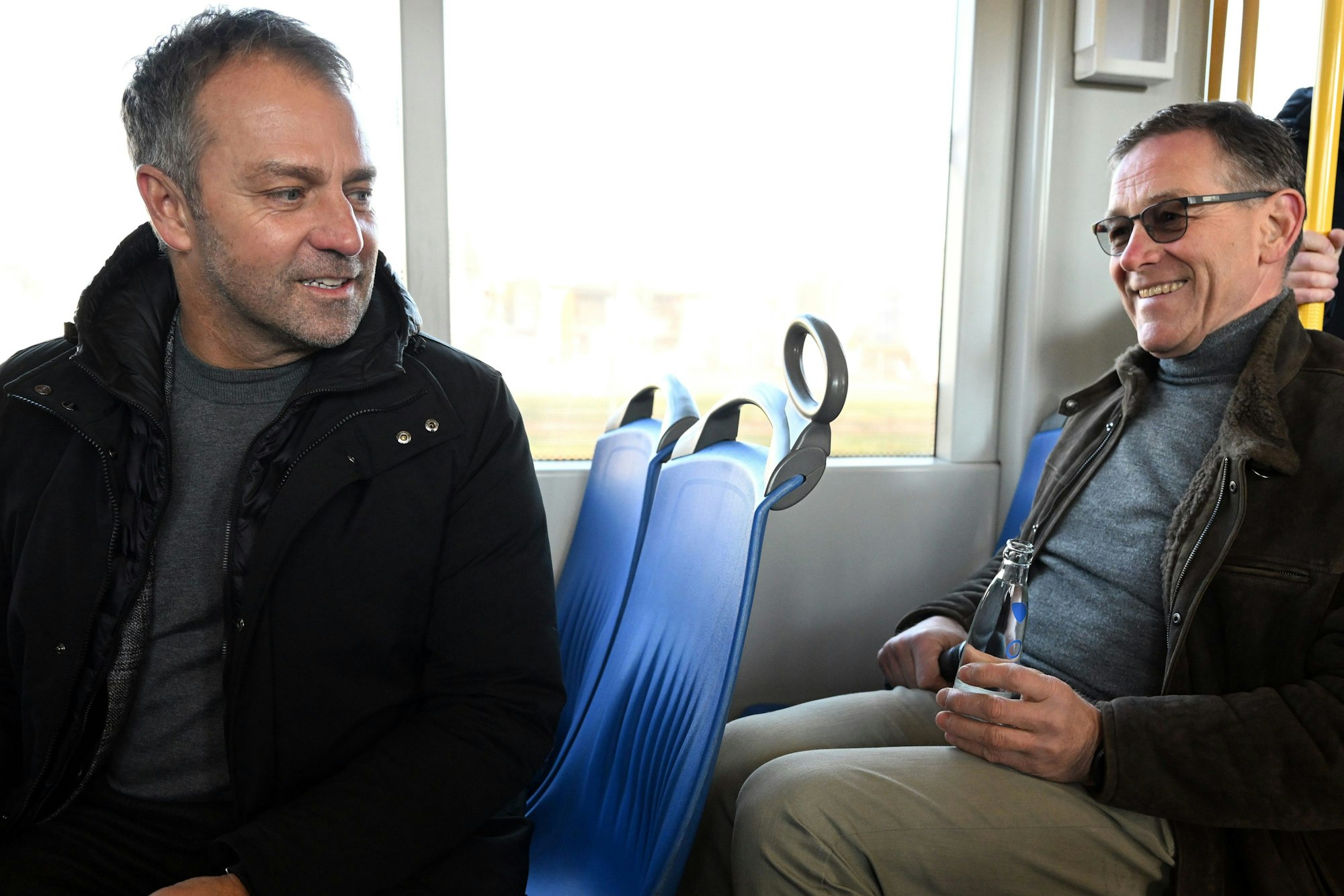 Fußball-Bundestrainer Hansi Flick und Handball-Bundestrainer Alfred Gislason sitzen zusammen in einer Straßenbahn.