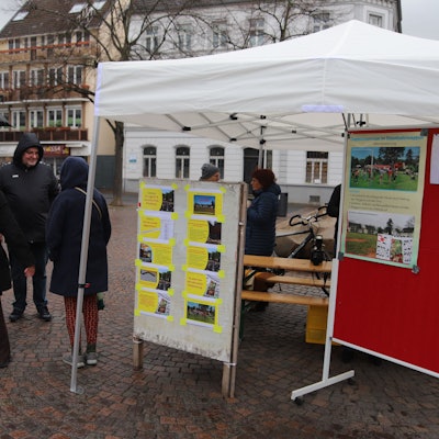 Blick auf das weiße Infostand-Zelt. Einige Leute stehen auf dem Platz davor.
