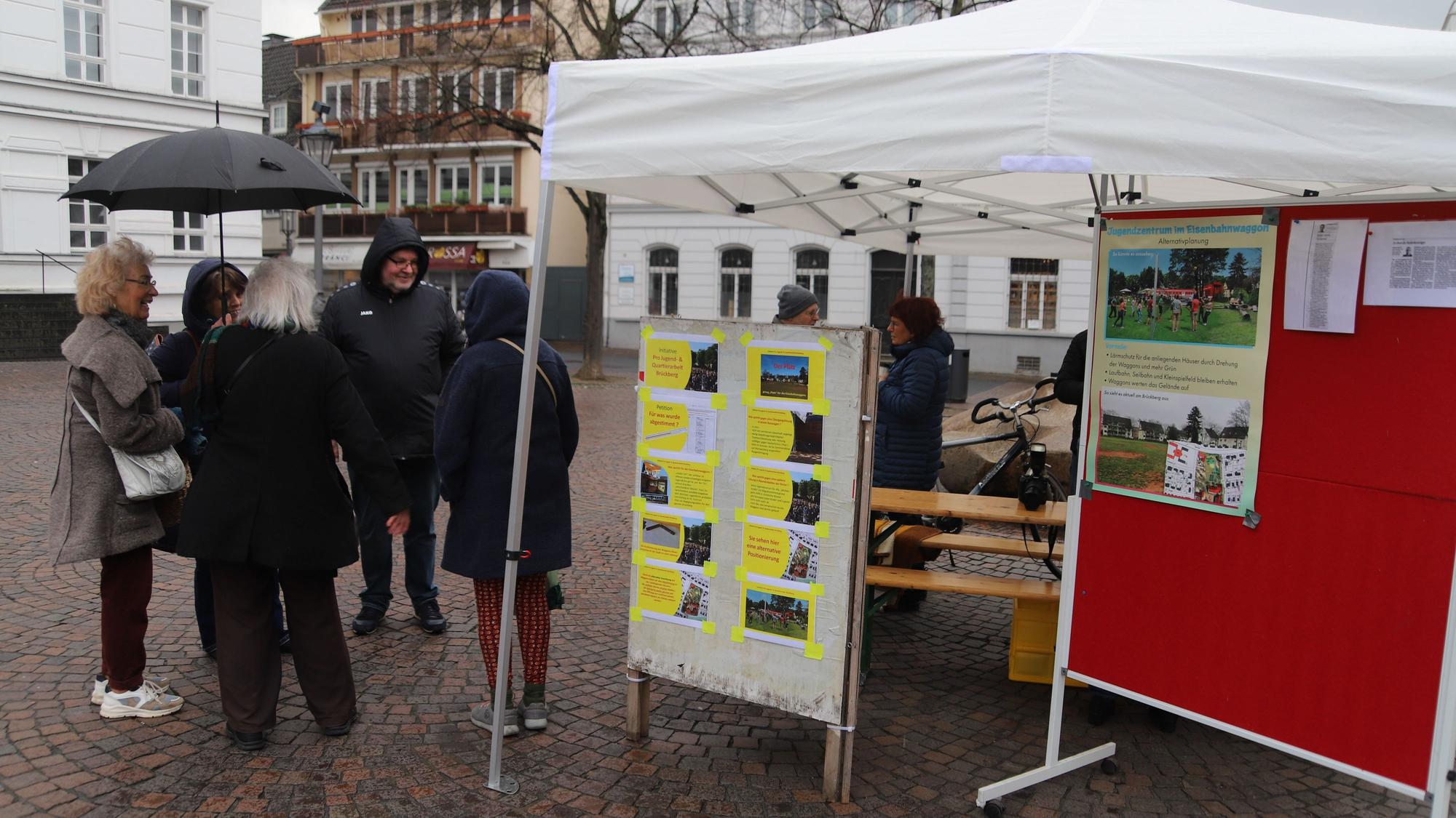 Blick auf das weiße Infostand-Zelt. Einige Leute stehen auf dem Platz davor.