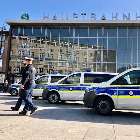 Drei Polizeiwagen stehen vor dem Kölner Hauptbahnhof. Zwei Polizisten stehen daneben.