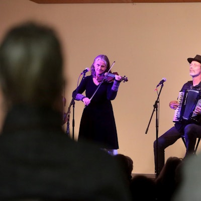 Eine Musikerin spielt Geige, ein Musiker neben ihr Akkordeon. Im Vordergrund sind schemenhaft zwei Zuschauer von hinten zu sehen.