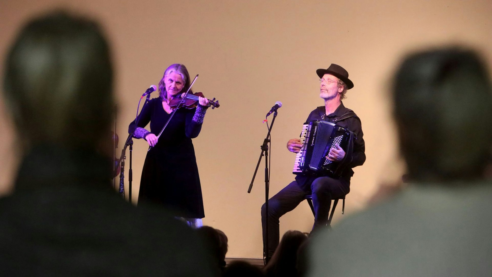 Eine Musikerin spielt Geige, ein Musiker neben ihr Akkordeon. Im Vordergrund sind schemenhaft zwei Zuschauer von hinten zu sehen.