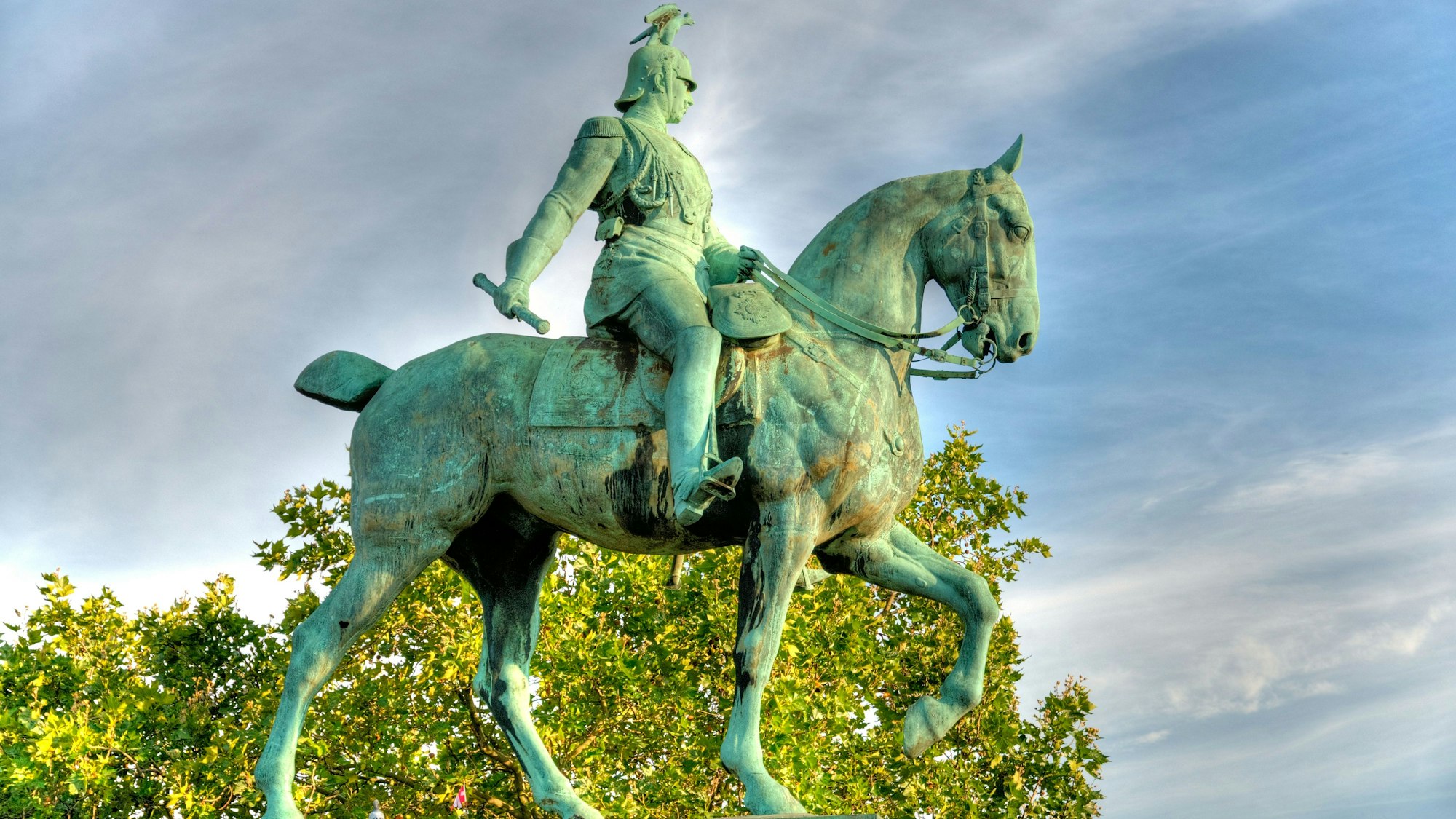 Vor blaugrauem Himmel und grünem Laub heben sich ein kraftvoll voranschreitendes Pferd und sein entschlossen wirkender Reiter Kaiser Wilhelm II. ab. Der Kaiser hält die Zügel in der linken Hand, in der rechten, nach hinten gestreckten Hand hält er einen Stab oder eine Schriftrolle. Der Kaiser blickt von seiner erhobenen Position aus links am Pferd vorbei, vom Betrachter weg.