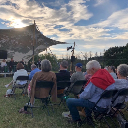 Personen sitzen auf Klappstühlen auf einer Wiese im Sonnenuntergang, mit Blick auf eine Bühne unter einem Sonnensegel.
