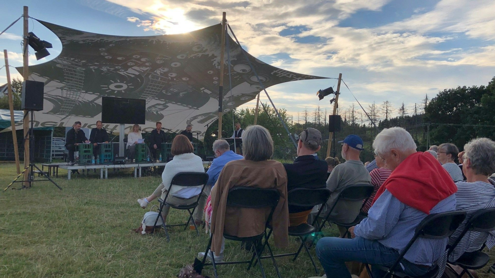Personen sitzen auf Klappstühlen auf einer Wiese im Sonnenuntergang, mit Blick auf eine Bühne unter einem Sonnensegel.