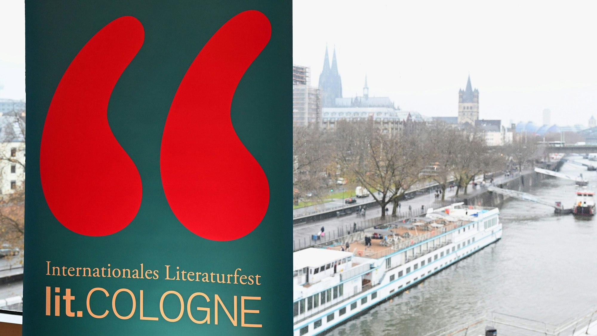 Ein Schild mit dem Logo der lit. Cologne am Rheinufer mit Blick auf den Dom.