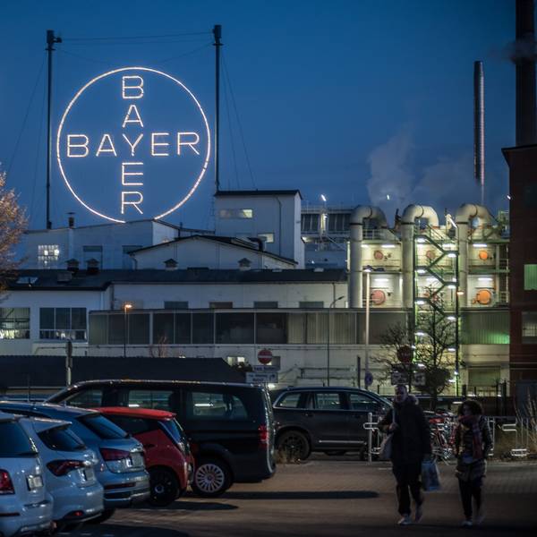 Das leuchtende Bayer-Kreuz ist aus einiger Entfernung vor parkenden Autos im Chempark Leverkusen abgebildet. Schornsteine qualmen im rechten Teil des Bildes, links befinden sich zwei Bäume.