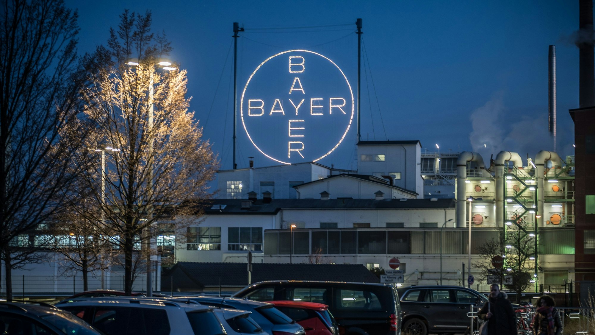 Das leuchtende Bayer-Kreuz ist aus einiger Entfernung vor parkenden Autos im Chempark Leverkusen abgebildet. Schornsteine qualmen im rechten Teil des Bildes, links befinden sich zwei Bäume.
