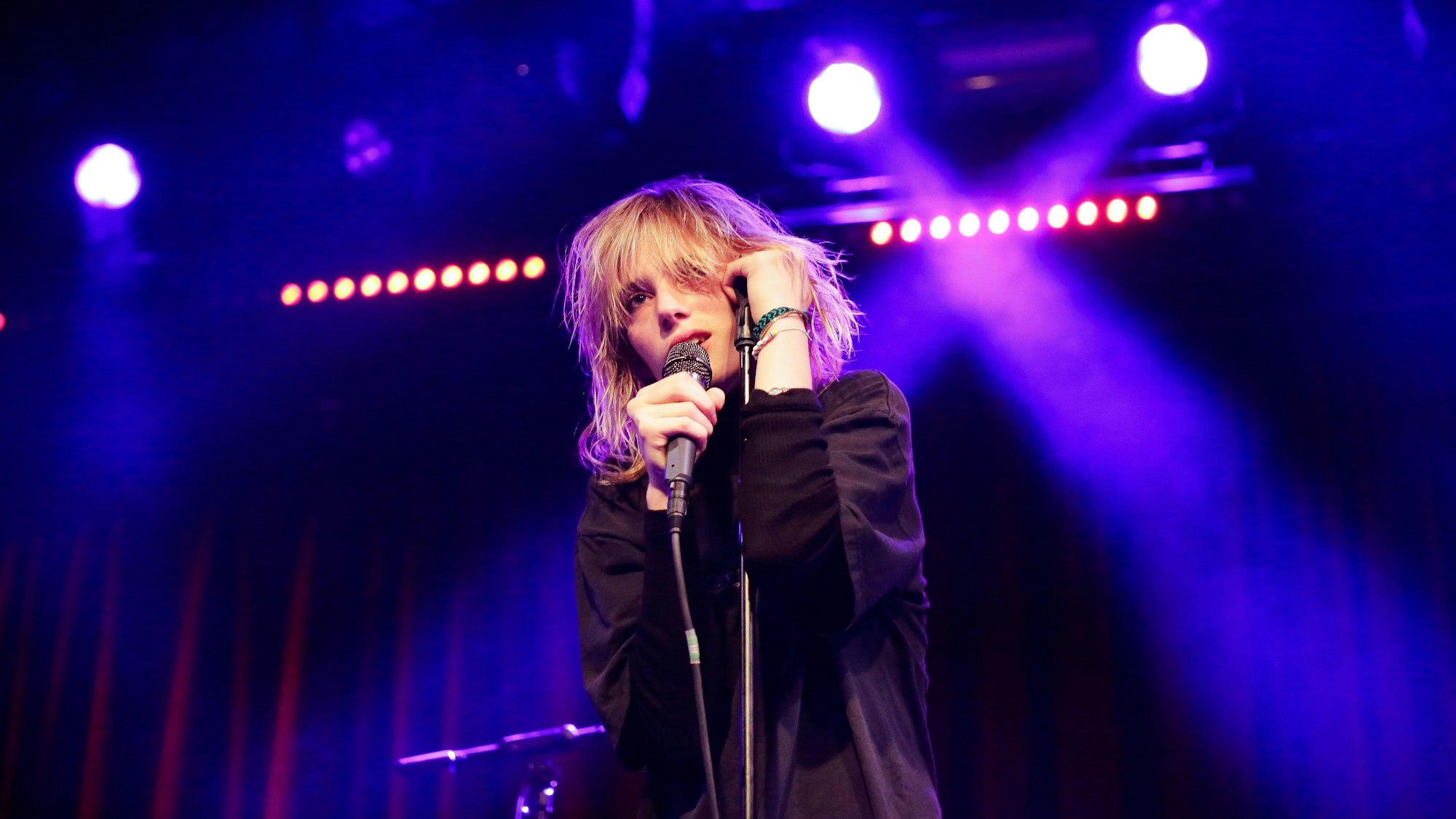 Maya Hawke singt auf der Bühne der Kantine in Köln. In einer Hand hält sie das Mikrofon, mit der anderen stützt sie sich auf den Mikrofonständer.
