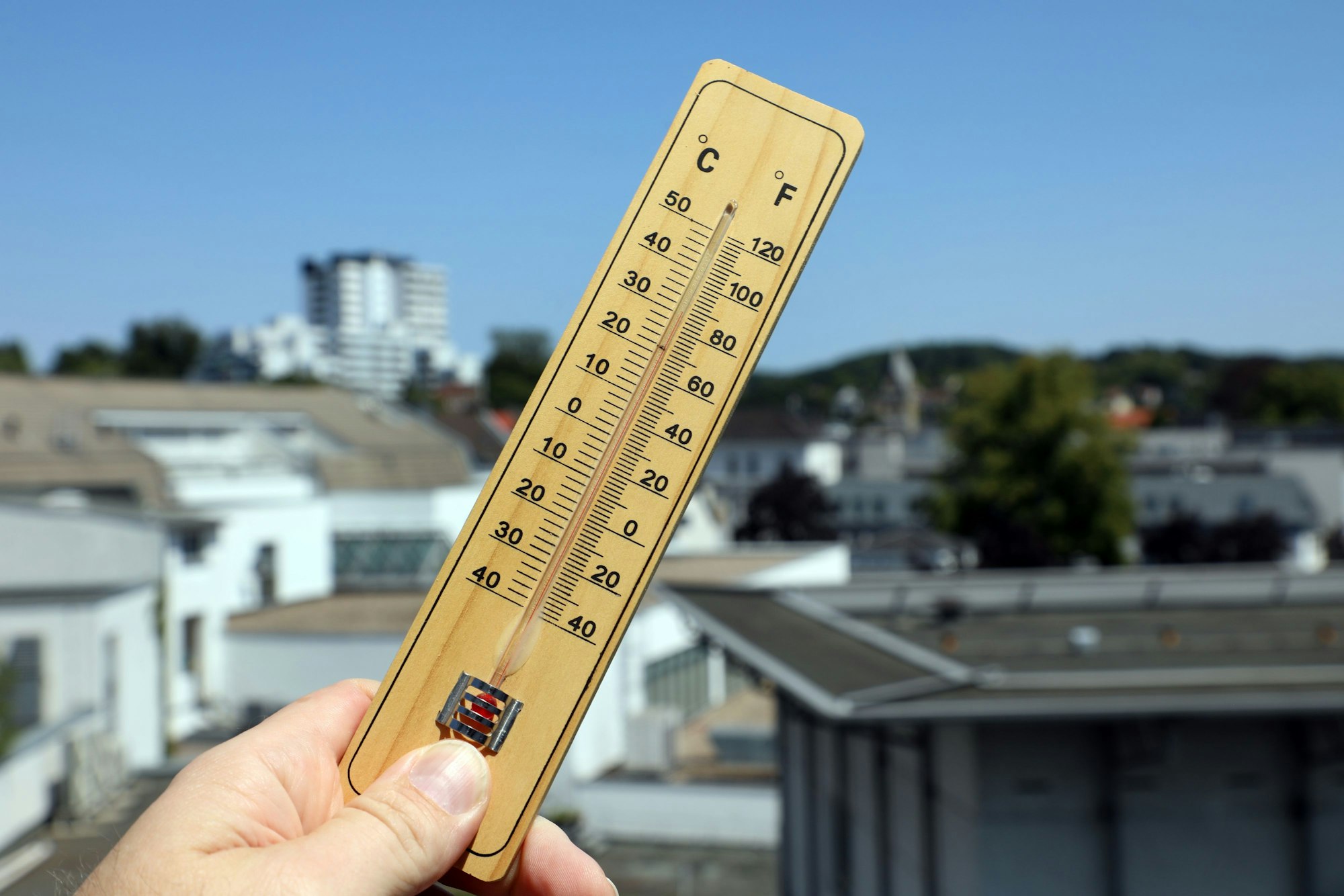 Jemand hält ein Thermometer in Bergisch Gladbach vor den strahlend blauen Himmel und die Häuserkulisse im Hintergrund.