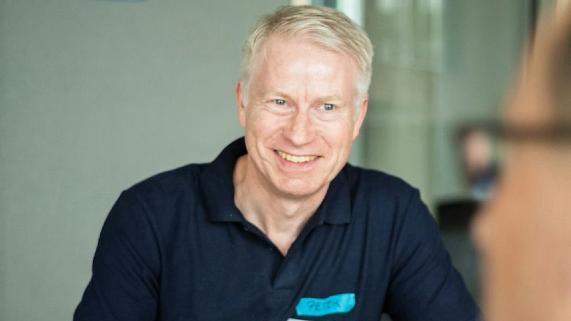 Peter Muth trägt ein blaues T-Shirt und lächelt in die Kamera.