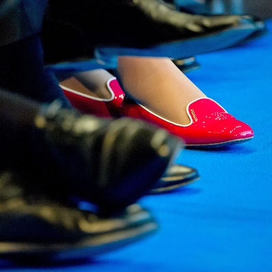 In roten Schuhen sitzt eine Teilnehmerin zwischen Männern in dunklen Anzügen und passendem Schuhwerk.