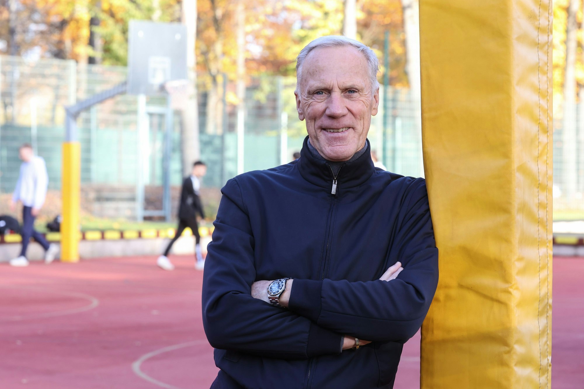 Prof. Dr. Ingo Froböse,
Universitätsprofessor für Prävention und Rehabilitation im Sport an der Deutschen Sporthochschule Köln, steht mit verschränkten Armen an einen Pfosten gelehnt auf einem Basketballplatz. Er lächelt in die Kamera. Im Hintergrund sieht man verschwommen zwei Sportler über den Platz gehen.