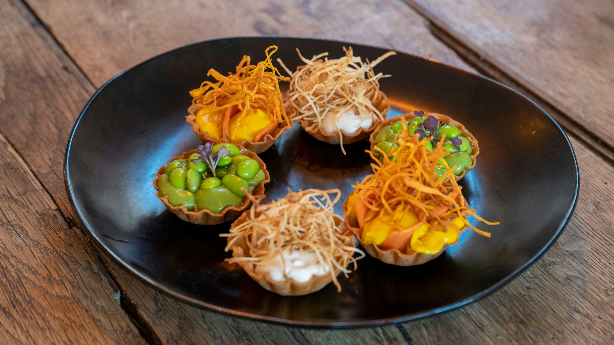 Tartelettes mit Edamame, Karotten und Sellerie stehen auf einem schwarzen Teller.