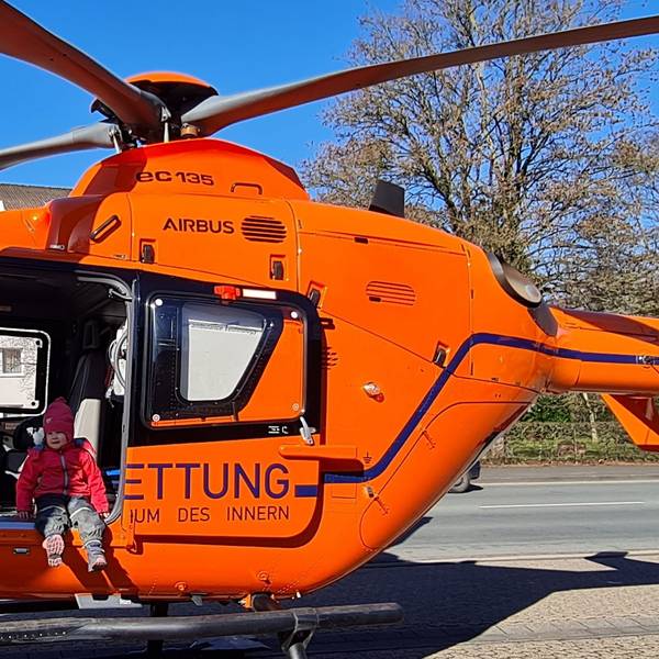 Ein Kind in Matschhose und mit Mütze sitzt mit baumelnden Beinen in der offenen Tür eines Helikopters der Luftrettung.