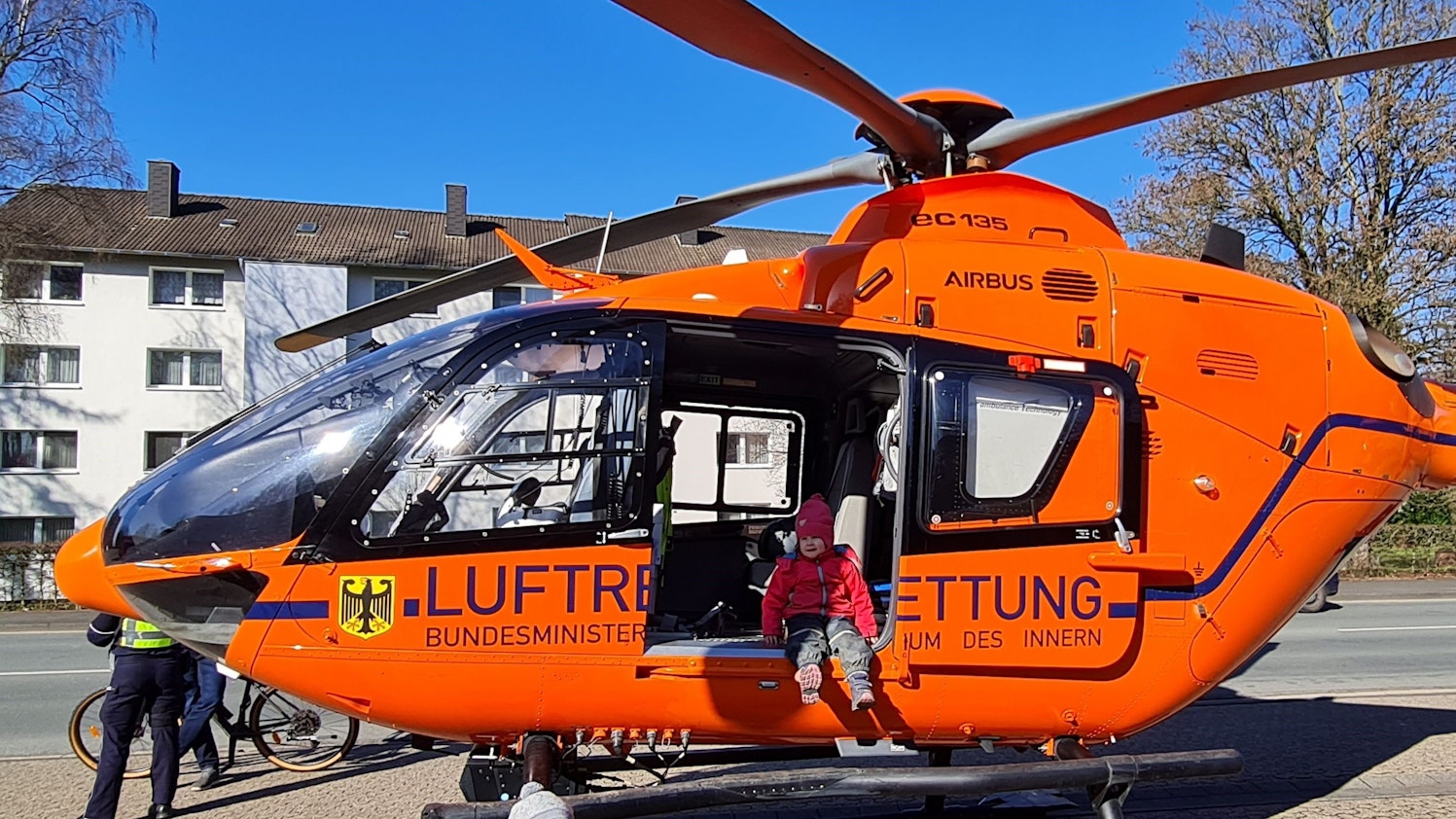 Ein Kind in Matschhose und mit Mütze sitzt mit baumelnden Beinen in der offenen Tür eines Helikopters der Luftrettung.