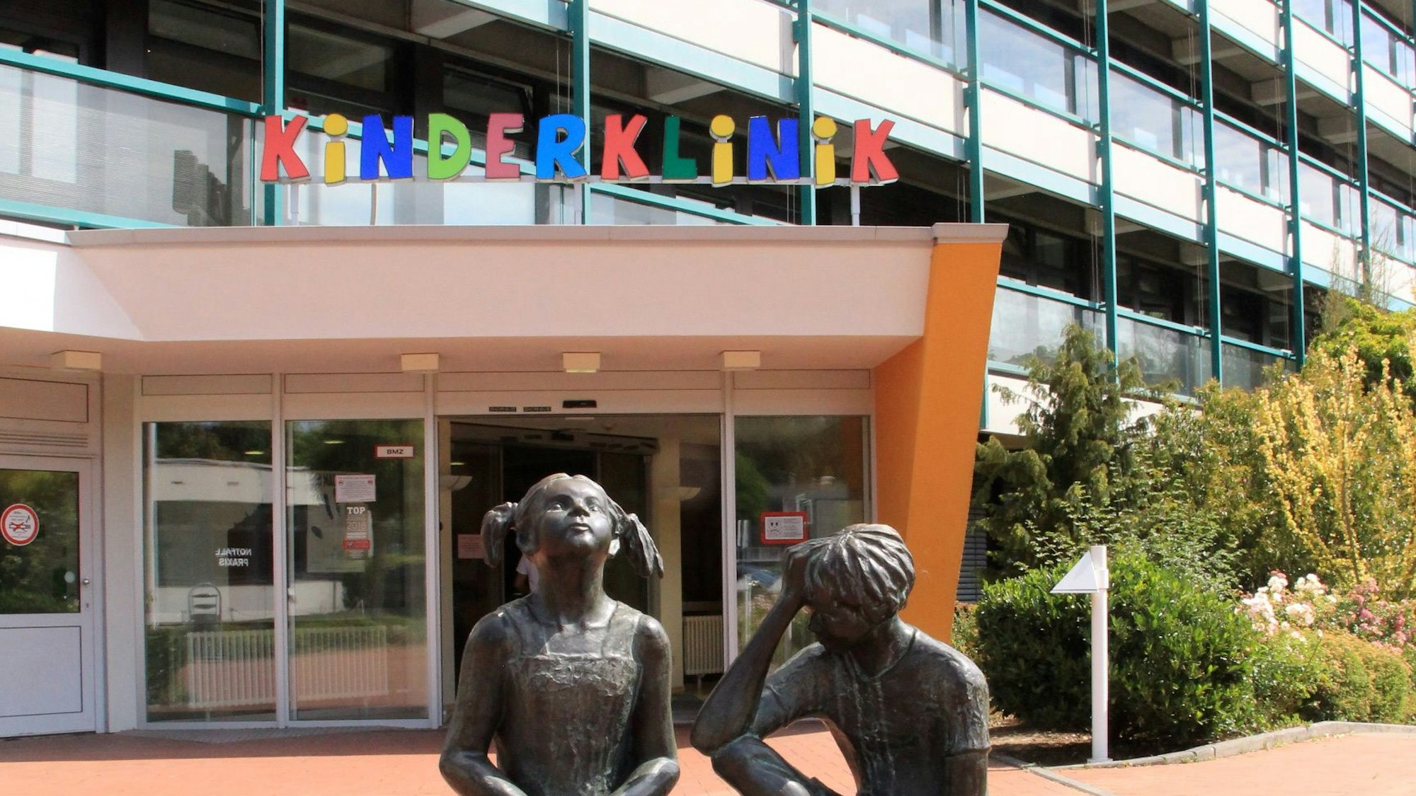 Über dem Eingang steht in bunten Buchstaben Kinderklinik. Im Vordergrund sind zwei Statuen von Kindern zu sehen.
